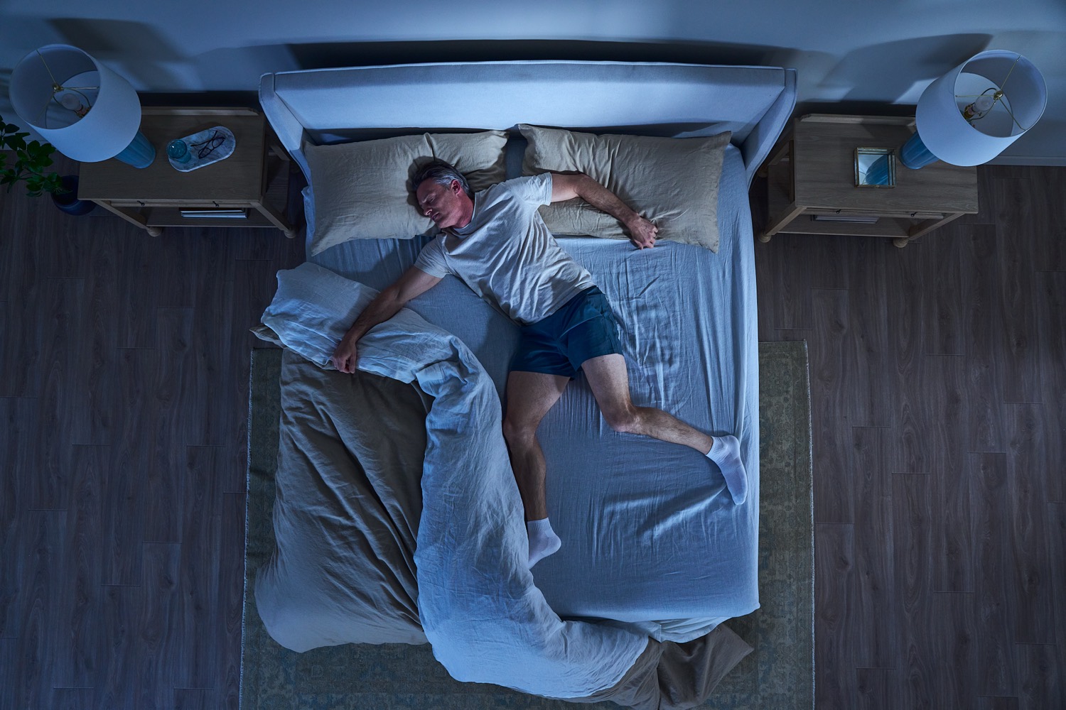 Overhead view of a man sprawled restlessly across the bed at night, covers kicked aside.